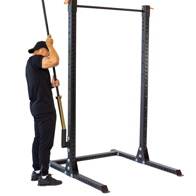 A person in black clothing and a cap removes a barbell from the Bells of Steel Vertical Mount Barbell Holder Rack Attachment and inserts it into a landmine attachment on a squat rack to prepare for strength training.