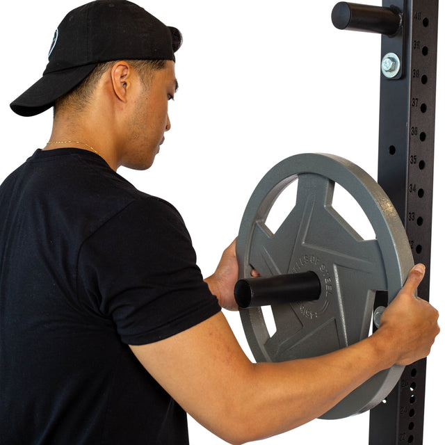 A person in a black shirt and cap is loading Olympic weight plates onto a power rack using Bells of Steel Bolt-On Plate Pegs in a gym.