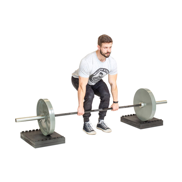 A man in a gray t-shirt and black pants sets up for deficit deadlifts on Bells of Steel Stackable Pull Blocks, standing on rubber mats with a barbell loaded with large plates, against a white background.
