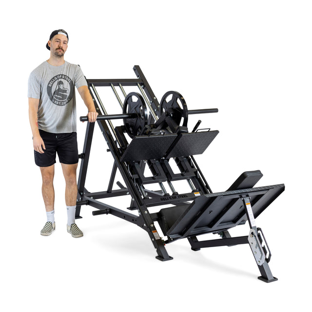 A man in a gray t-shirt, black shorts, and a backward cap stands next to the Bells of Steel 2-in-1 Iso Leg Press & Hack Squat Machine (The Juggernaut) with weight plates loaded.