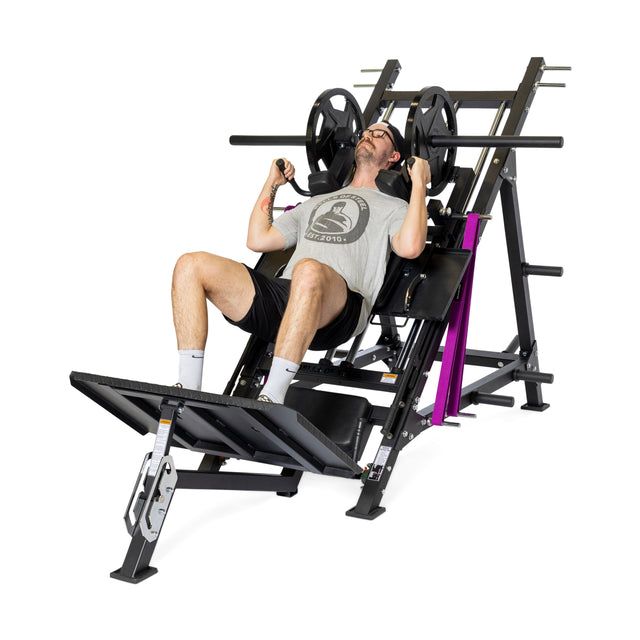 A man in a gray t-shirt, black shorts, glasses, and white socks uses the Bells of Steel 2-in-1 Iso Leg Press & Hack Squat Machine (The Juggernaut), seated and pushing the footplate while gripping the handles in a gym.