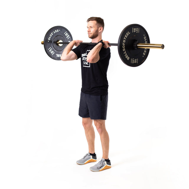 A man stands against a plain white background, holding a Bells of Steel Olympic Weightlifting Barbell at shoulder height in the front rack position, dressed in a black t-shirt, black shorts, and gray sneakers, preparing to lift.