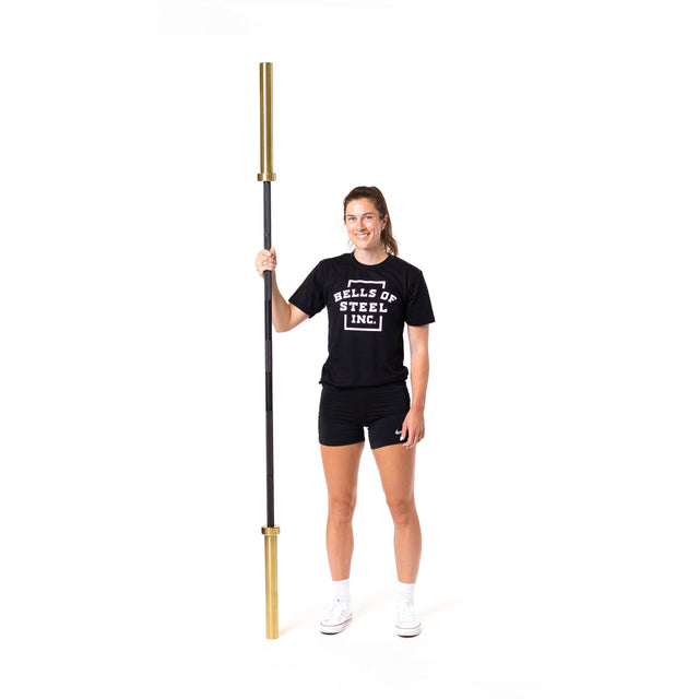 A woman in athletic wear smiles while holding a Bells of Steel Olympic Weightlifting Barbell vertically, with the brand name on her black T-shirt, against a plain white background.