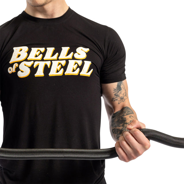A tattooed person in a black Bells of Steel t-shirt holds a coiled battle rope, ready for arm exercises like bicep curls using the Bells of Steel Rackable EZ Curl Bar, all set against a plain white background.