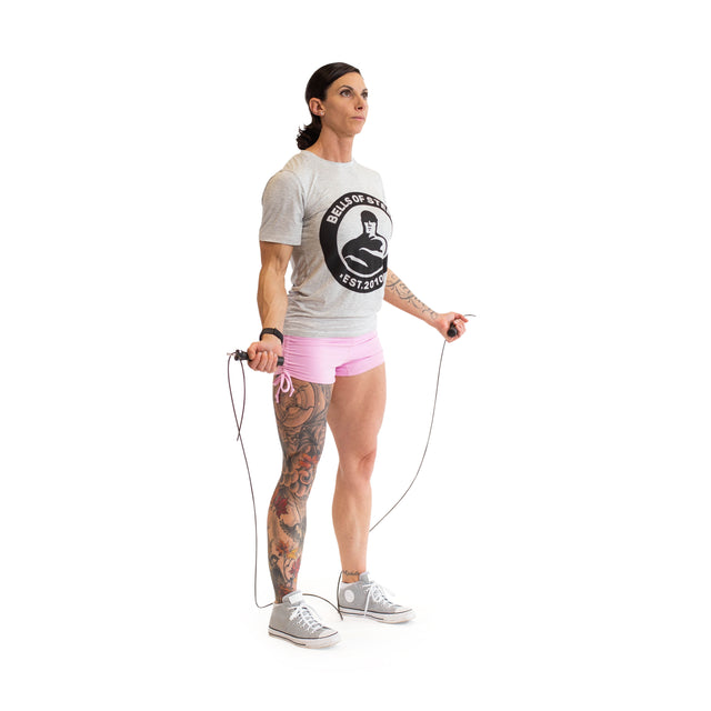 On a white background, a person holds the Bells of Steel Jump Rope with ergonomic handles, wearing a gray logo T-shirt, pink shorts, and gray sneakers. Tattoos adorn one leg. Focused and ready for an intense cardio session, they prepare to jump.