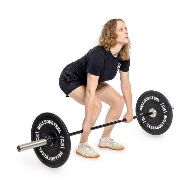 A woman in a black outfit, in a squat stance on a white background, prepares to lift the Bells of Steel Juno Bar – Women’s Utility Bar, loaded with black weight plates.
