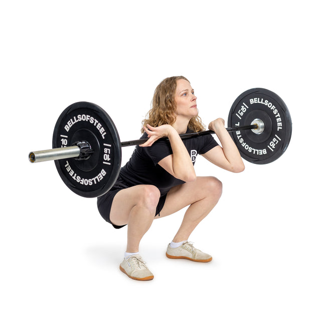A woman with curly hair wearing a black shirt and shorts performs a deep front squat with the Bells of Steel Juno Bar - Women’s Utility Bar (25mm) resting on her shoulders. The background is white.