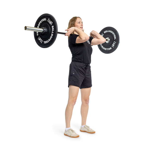 A person in a black shirt and shorts lifts the Bells of Steel Juno Bar - Women’s Utility Bar with black weight plates labeled "Bells of Steel" in a front rack position, standing on a white background.