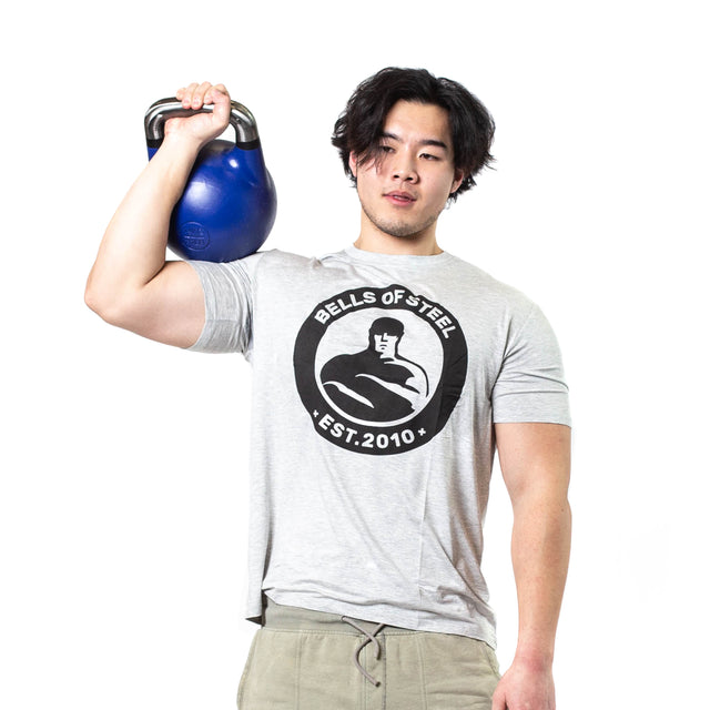 A man in a light gray "Bells of Steel EST. 2010" t-shirt and khaki pants stands against a white background, holding a blue Bells of Steel Competition Kettlebell with an ergonomic design on his shoulder.