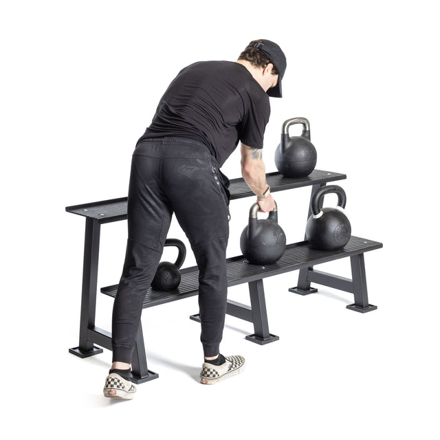 A person in black athletic wear and checkered shoes is reaching for a competition kettlebell on the Bells of Steel Kettlebell Rack, which holds multiple kettlebells and fits perfectly in any modern home gym, set against a white background.