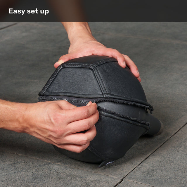 Hands zip a black, polygonal leather Bells of Steel USA Kettlebell Wrap over an object on a gray stone floor beneath the text “Easy set up.”.