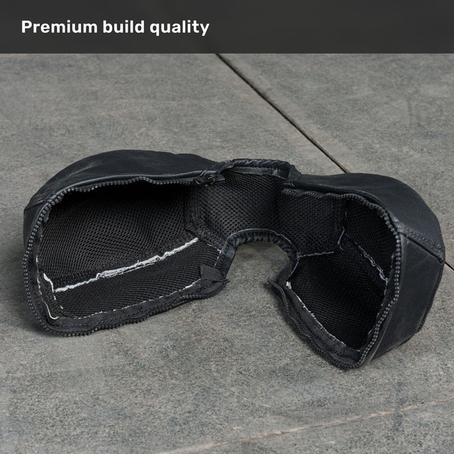 A close-up of the Bells of Steel USA Kettlebell Wrap, a black padded fabric accessory with visible stitching, resting on a gray textured surface. At the top, the image features the text “Premium build quality,” emphasizing the exceptional craftsmanship of this wrap designed specifically for use with Competition Kettlebells.