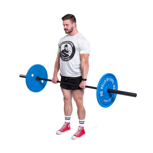 A man in a gray T-shirt, black shorts, and red sneakers holds a barbell with Bells of Steel Calibrated Powerlifting Plates - KG. Standing on a white background, he looks focused and strong, showcasing his strength with these color-coded plates of standardized diameters.