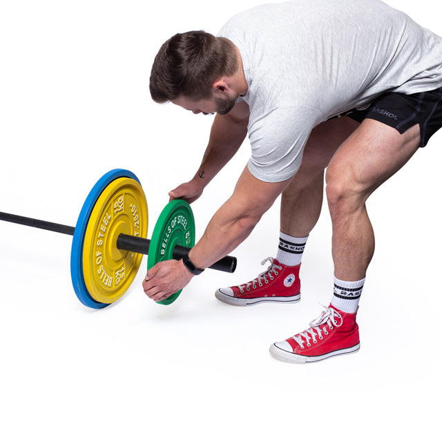 A man in athletic wear, wearing red high-top sneakers, bends over to add a green Calibrated Powerlifting Plate from Bells of Steel to a barbell already loaded with yellow and blue weights, standing against a white backdrop.