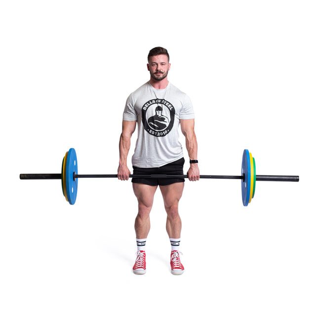 A bearded man wearing a gray T-shirt, black shorts, red shoes, and socks holds a barbell with Bells of Steel's Calibrated Powerlifting Plates - KG Sets. These colorful plates meet IPF specs. The background is plain white.