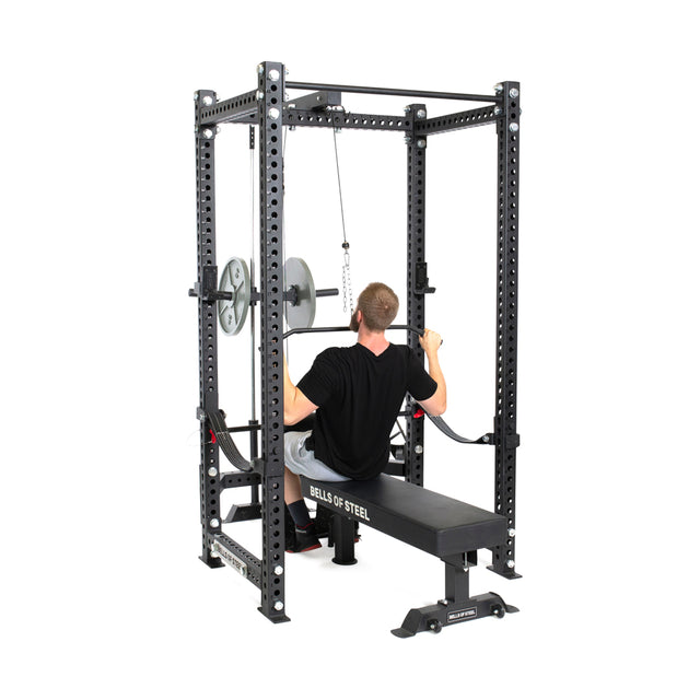 A man wearing a black shirt uses the Bells of Steel power rack, featuring the Lat Pulldown & Low Row Rack Attachment - Hydra & Manticore. He pulls the cable while seated on a bench, loaded with two Olympic plates per side.