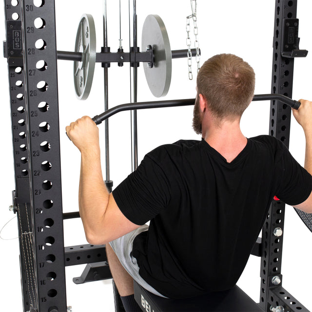 A bearded man in a black T-shirt and gray shorts uses the Bells of Steel "Lat Pulldown & Low Row Rack Attachment - Hydra & Manticore" as he concentrates on cable exercises with Olympic weight plates stacked on the gym machine.