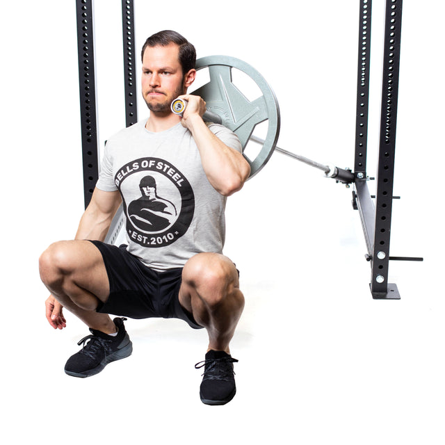A man squats in the gym with a barbell on his shoulders, showcasing a strength workout. He's in a gray "Bells of Steel" T-shirt, black shorts, and shoes against a white backdrop, highlighting his focus and equipment. Behind him is the innovative Bells of Steel Landmine Rack Attachment.