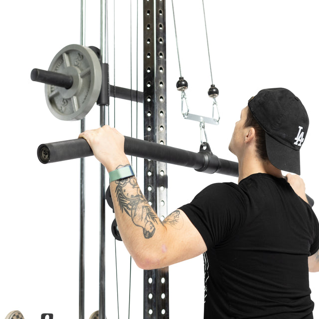 Wearing a black t-shirt and backward cap, a person uses the Bells of Steel Fat Bar - Lat Pulldown Bar, gripping the cable machine attachment with both hands. A tattoo on their left forearm highlights focused grip strength.