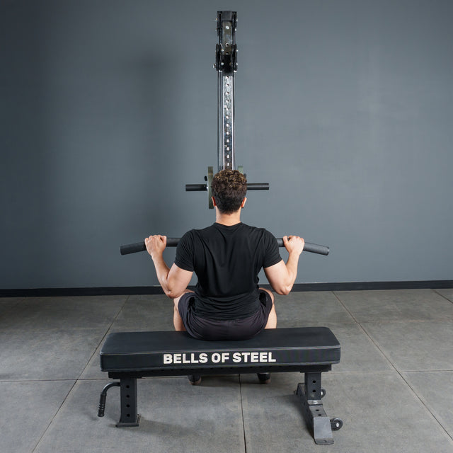 In a gym, a person on a Bells of Steel bench faces a cable machine and grasps the Bells of Steel Fat Bar - Lat Pulldown Bar, ready to perform lat pulldowns to increase grip strength.