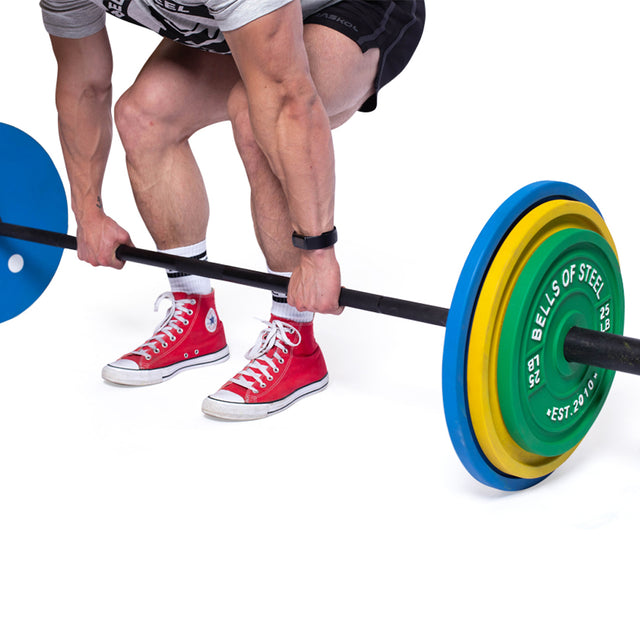 Wearing red high-top sneakers and a gray shirt, a person stands in a squat stance preparing to lift a barbell loaded with Bells of Steel's Calibrated Powerlifting Plates - LB in blue, yellow, and green against a white backdrop, highlighting their powerlifting technique.