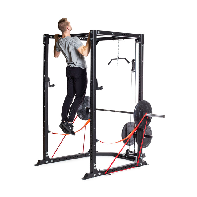 A man in a gray t-shirt and black pants does a pull-up on the Bells of Steel Light Commercial Power Rack (2.3" x 2.3", ⅝" holes), equipped with weights, resistance bands, and barbell protection, against a plain white background.