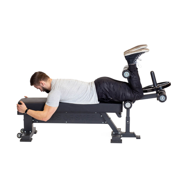 A man in a gray shirt, black pants, and sneakers uses the Bells of Steel Bench Attachments to perform a hamstring curl on a bench, gripping the handles. He is positioned face down against a white background.