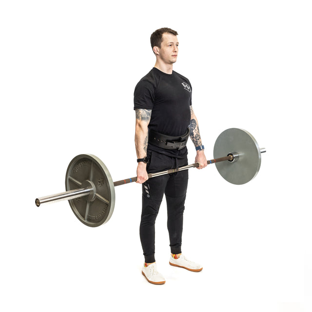 A man in athletic clothing stands upright holding a loaded barbell at thigh level, indicating the end of a deadlift, while wearing the Bells of Steel Lever Belt against a plain white background.