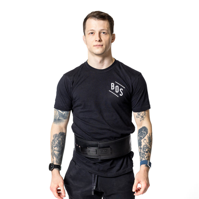 A man with short brown hair, black "BOS" t-shirt, black athletic pants, Bells of Steel Lever Belt, and smartwatch stands facing forward. He has tattoos on both arms, a neutral expression, and a white background.