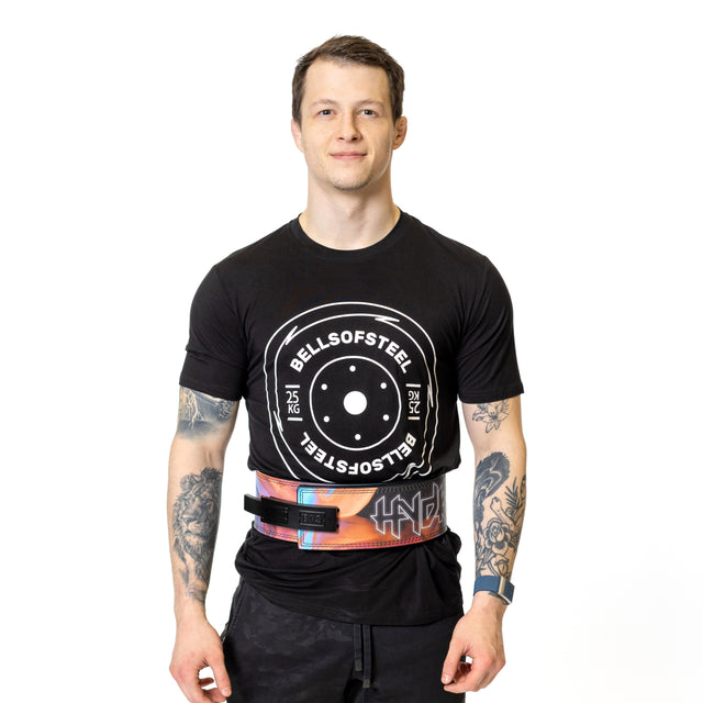 A man with short brown hair, tattoos, and a smartwatch stands smiling against a white background, wearing a black Bells of Steel t-shirt and shorts, with the colorful Bells of Steel Lever Belt from his waist.