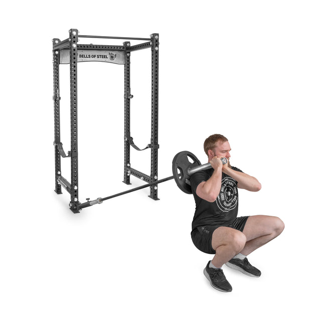 A person, in athletic attire, performs a front squat with an Olympic barbell across their shoulders. In the background, a Bells of Steel squat rack with a Landmine Rack Attachment is situated on a plain floor, highlighting the strength workout.