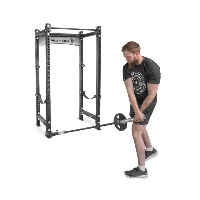 A person works out with a Bells of Steel Landmine Rack Attachment, skillfully managing an Olympic barbell in both hands for rotational moves. The rack displays the "Bells of Steel" brand, and they're wearing a dark shirt and shorts.