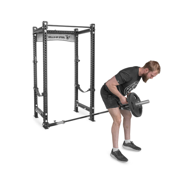 A man performs a T-bar row using an Olympic barbell with weight plates attached to a Bells of Steel Landmine Rack Attachment. Bending forward in black t-shirt, shorts, and sneakers, he showcases his strength workouts beside the branded squat rack.