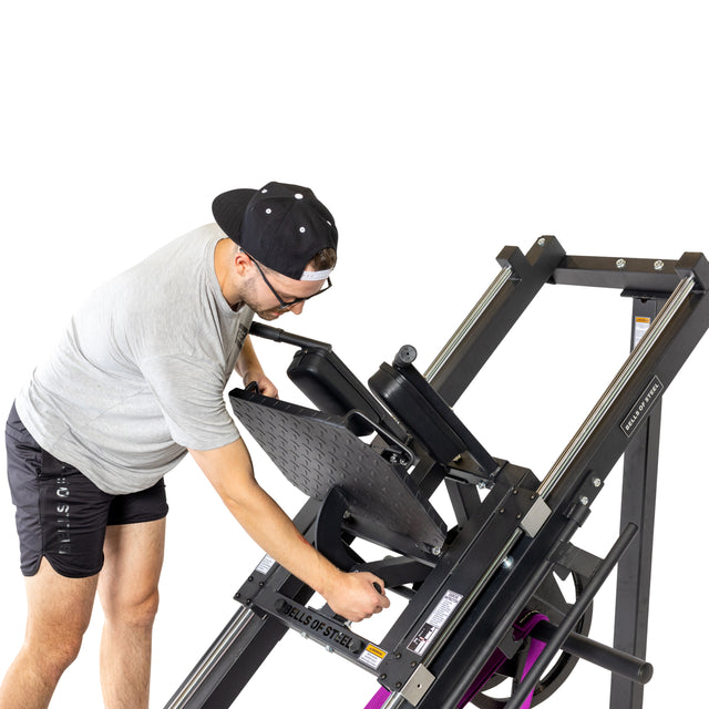 A man in a gray shirt, black shorts, and a backward cap adjusts the footplate on the Bells of Steel Leg Press Hack Squat Machine against a white background.