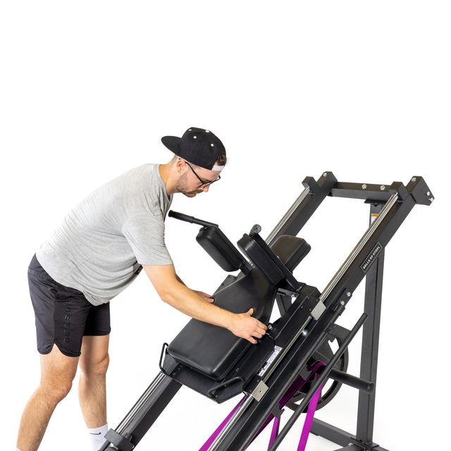 A man in a cap, glasses, gray shirt, and black shorts adjusts the Bells of Steel Leg Press Hack Squat Machine with purple resistance bands, perfect for any home gym, set against a white background.