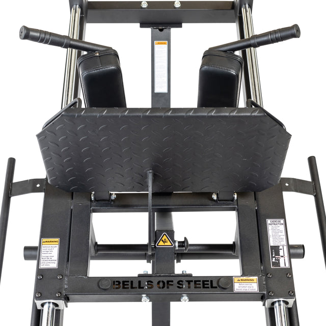 A top-down close-up of the Bells of Steel Leg Press Hack Squat Machine shows its black footplate, padded handles, and safety labels. The empty 2-in-1 machine is ideal for a home gym setup.