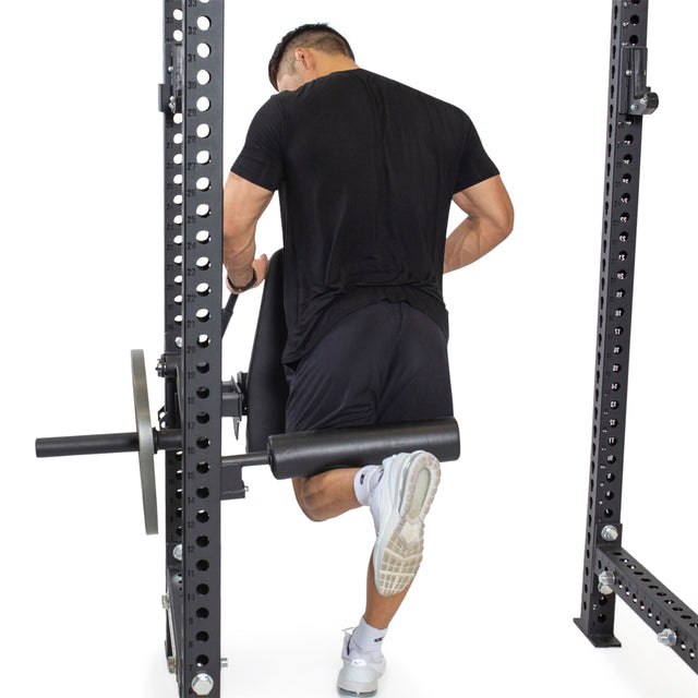 A man in black athletic wear does a single-leg squat in his home gym using the Bells of Steel Leg Curl / Leg Extension Rack Attachment, gripping the rack for balance with one leg bent behind the support.
