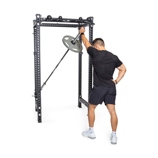 A man in athletic wear stands beside a Bells of Steel Manticore Four Post Power Rack - Prebuilt (3" x 3", 1" Holes), gripping a barbell with weight plate, preparing for a landmine exercise in his home gym, back to the camera and one hand on his hip.