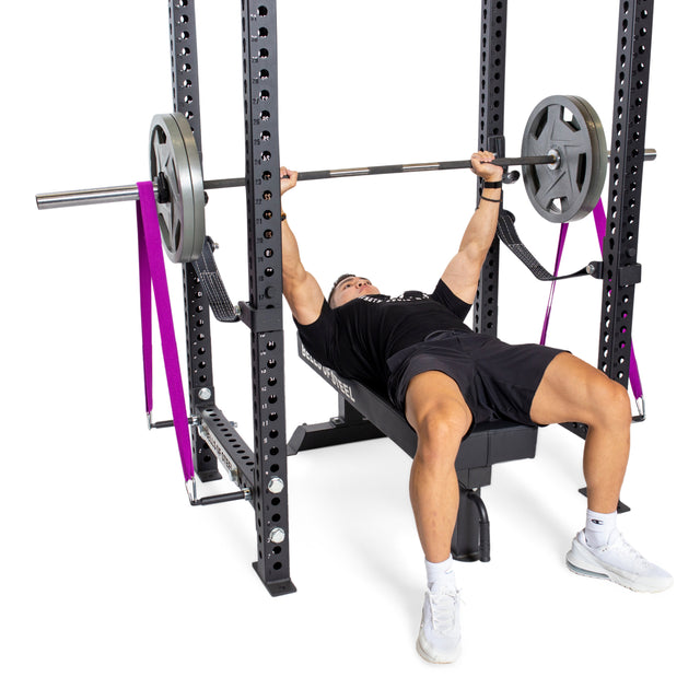 A person bench presses in their home gym using the Bells of Steel Manticore Four Post Power Rack - Prebuilt (3" x 3", 1" Holes), lifting a barbell with weight plates and purple resistance bands on a weight bench.
