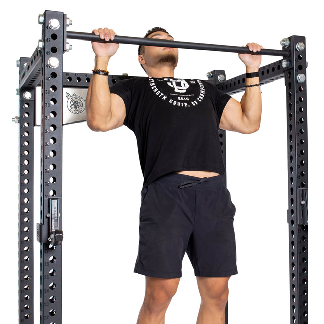 A man in a black t-shirt and shorts performs a pull-up on a metal bar attached to the Bells of Steel Manticore Four Post Power Rack - Prebuilt (3" x 3", 1" Holes), showcasing power and focus—ideal for any home gym.