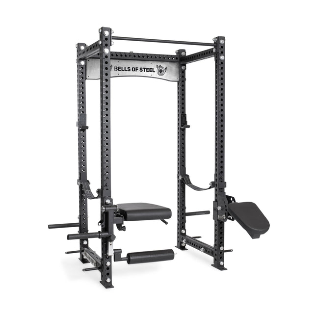 The Bells of Steel Manticore Four Post Power Rack - Prebuilt features black 3"x3" steel uprights, 1" holes, multiple attachments (bench, leg rollers, dip bars, angled foot platform), and a "Bells of Steel" label—ideal for any home gym.