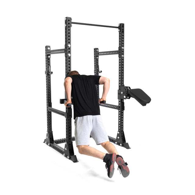 A person in a black shirt and gray shorts performs dips on parallel bars attached to the Bells of Steel Manticore Half Rack Builder (3" x 3", 1" Holes) with extra workout attachments, ideal for any home gym, against a white background.