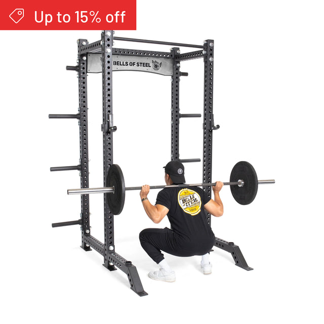 A person does a barbell squat inside a black Manticore Collegiate Power Rack Builder (3" x 3", 1" Holes) by Bells of Steel, wearing black clothes and a hat. "Up to 15% off" appears top left. Ideal for any home gym.