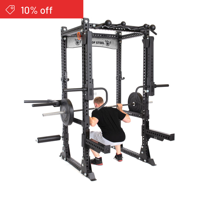 A man squats with a barbell inside the Bells of Steel Manticore Flat Foot Power Rack Builder (3" x 3", 1" Holes), equipped with attachments and weight plates—ideal for home gyms. A red banner in the corner reads "10% off.