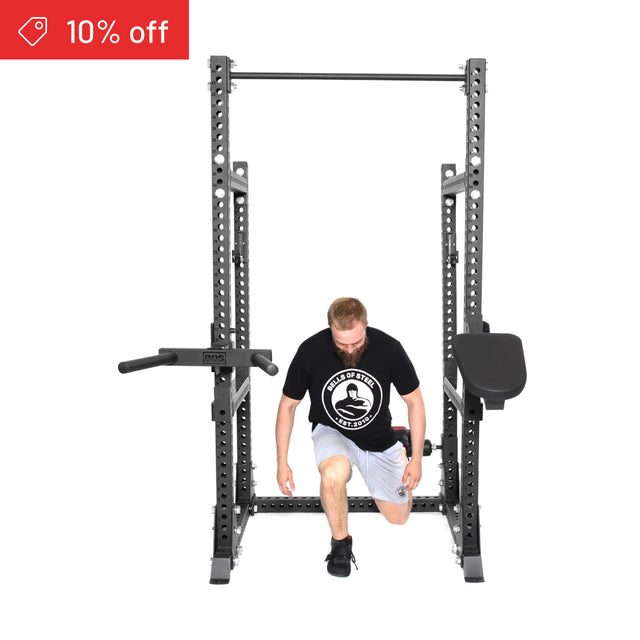 A man in athletic wear kneels inside the Bells of Steel Manticore Half Rack Builder (3" x 3", 1" Holes) with attachments. A red "10% off" label is shown on a white background—ideal for your home gym setup.