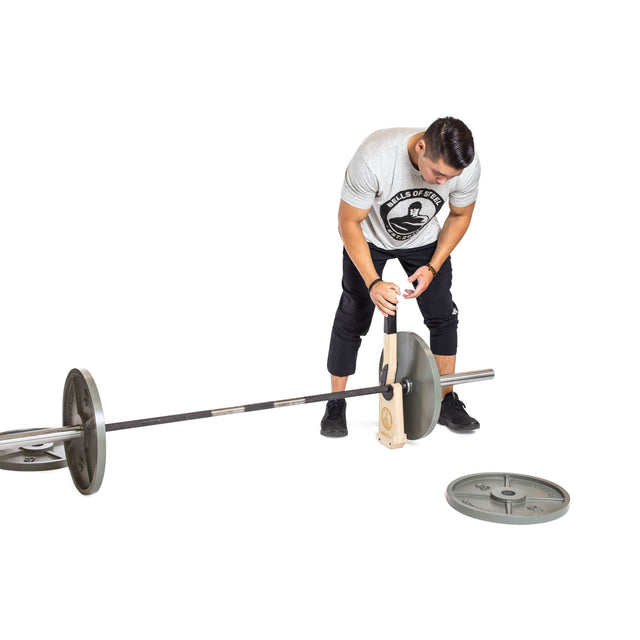 A man in a gray t-shirt uses the Bells of Steel Meat Hook Deadlift Jack to load a weight plate onto a barbell, with another plate resting on the floor nearby, all set against a white background.