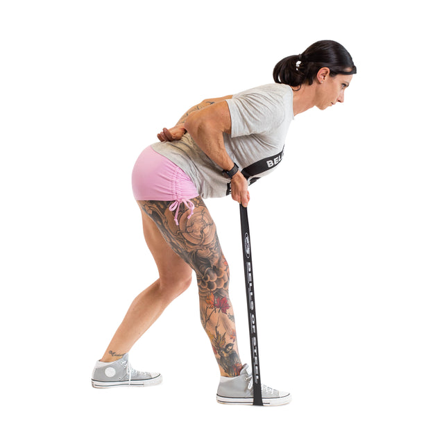 A woman in a gray shirt, pink shorts, and sneakers performs a bent-over row using Bells of Steel Mini Bands—one end under her foot and the other in her left hand—maintaining a flat back and slightly bent knees.