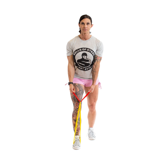 A tattooed woman faces forward, holding Bells of Steel Mini Bands anchored under her right foot—ideal versatile gym accessories. She wears a grey Bells of Steel T-shirt, pink shorts, and white sneakers against a white background.