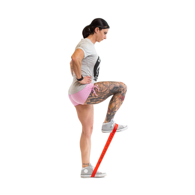 Wearing athletic wear, a woman performs a standing knee raise while pressing down on a Bells of Steel Mini Band, anchored under her left foot. She exercises against a plain white background.