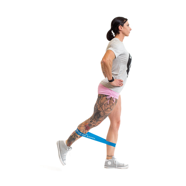 A person with tattoos on one leg uses Bells of Steel Mini Bands, wearing a gray t-shirt, pink shorts, and gray sneakers, extending one leg back with a blue band around their ankles against a white background.
